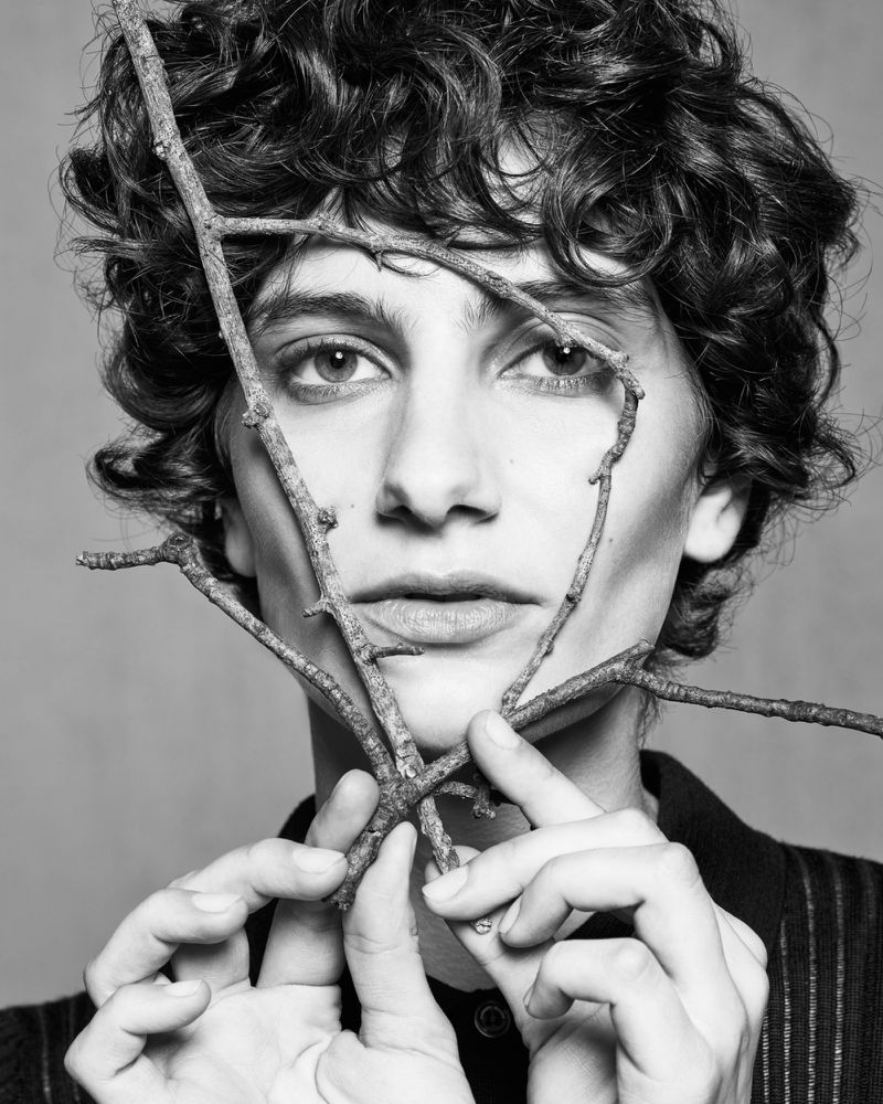 Monochromatic portrait of a young adult with curly dark hair, holding bare tree branches to frame the face and add artistic texture.