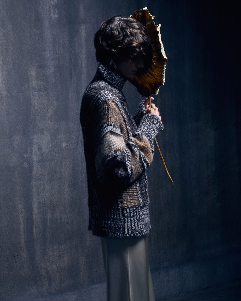 A person stands in front of a textured wall, wearing a textured sweater and holding a leaf in front of their face in a studio.