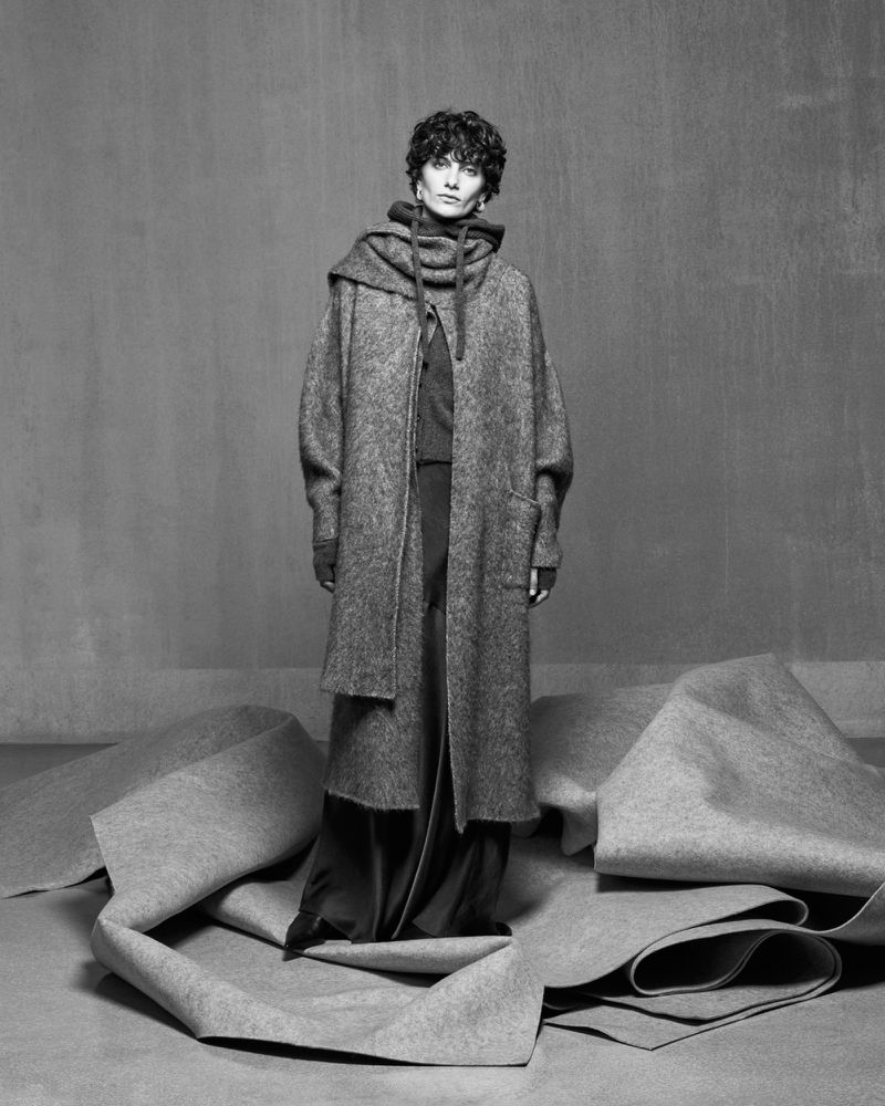 Model in an oversized grey knit coat and matching scarf standing on sculptural rolls of felt fabric against a grey textured studio backdrop.