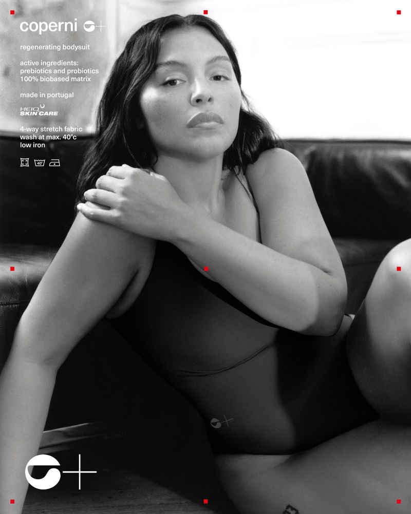 Adult woman poses in black bodysuit on a black couch in a studio. The advertisement features text from the brand Coperni. 