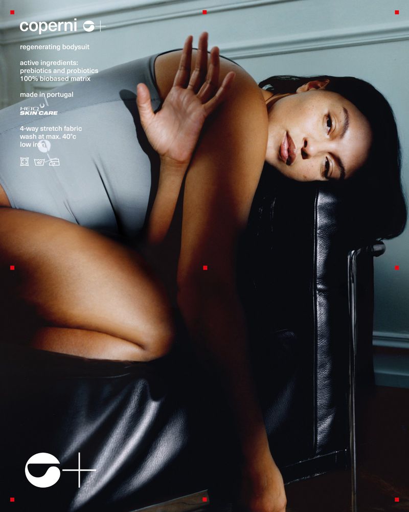A woman poses in a grey bodysuit on a black chair with copy of product information on a white wall; dark shadows and natural light.