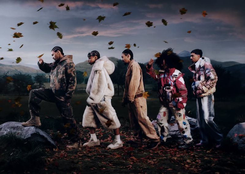 A group of five people walk on a path in a field under a cloudy sky with autumn leaves falling. They wear fashionable fall-themed clothing.