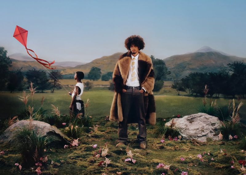 Man in a fur coat and a child with a kite in a scenic outdoor setting with hills and rocks, creating a high fashion editorial.