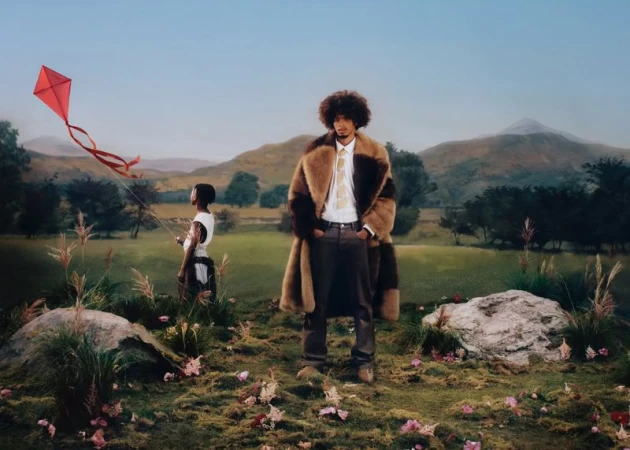 Man in a fur coat and a child with a kite in a scenic outdoor setting with hills and rocks, creating a high fashion editorial.