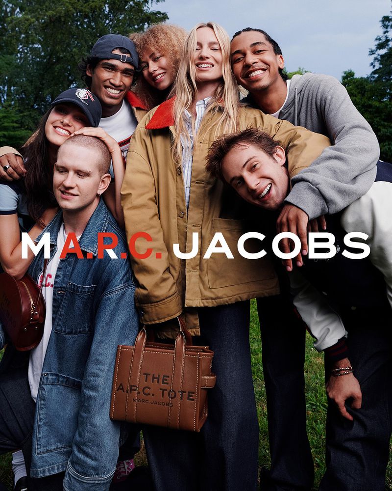 Group of seven smiling, multi-ethnic young adults gathered closely, posing with a brown leather Marc Jacobs tote bag in an outdoor setting.