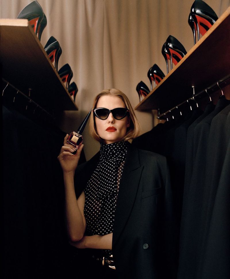 Blond woman in sunglasses and blazer holding perfume bottle in closet with black heels and clothing, dark and moody.