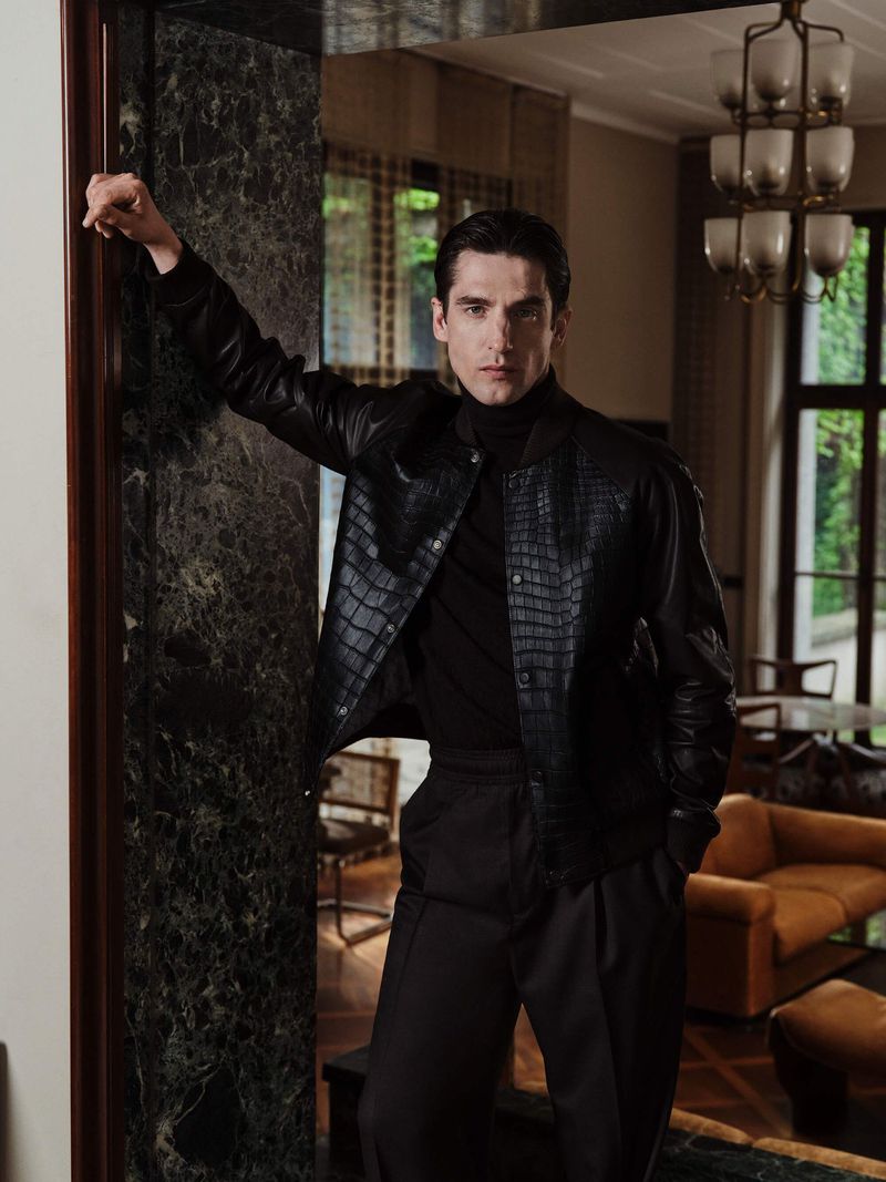A well-dressed adult male leans in a marble doorway, wearing a black leather jacket and dark slacks, in an elegant interior setting.