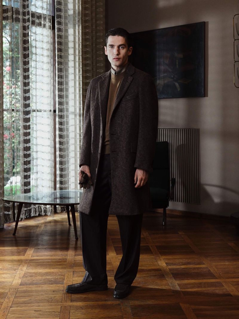 Full-length portrait of a dark-haired male model in a brown overcoat and black pants in a minimalist interior with parquet flooring.
