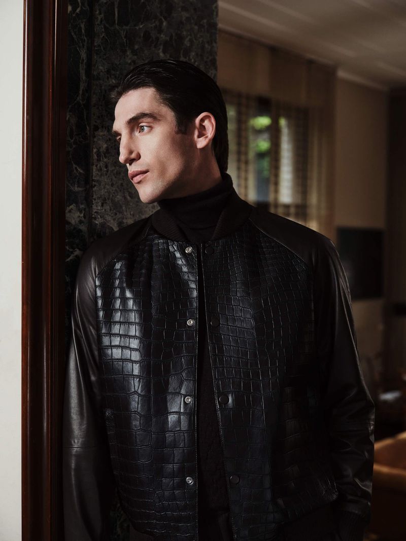 Elegant adult man in a textured black leather jacket stands indoors next to a marble wall looking left with soft window lighting.