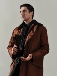 Man wearing brown coat and dark shirt holding leather gloves against a light grey background