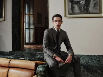 Model Anatol Modzelewski wearing a tailored grey suit and glasses seated on a green marble ledge in an elegant interior.