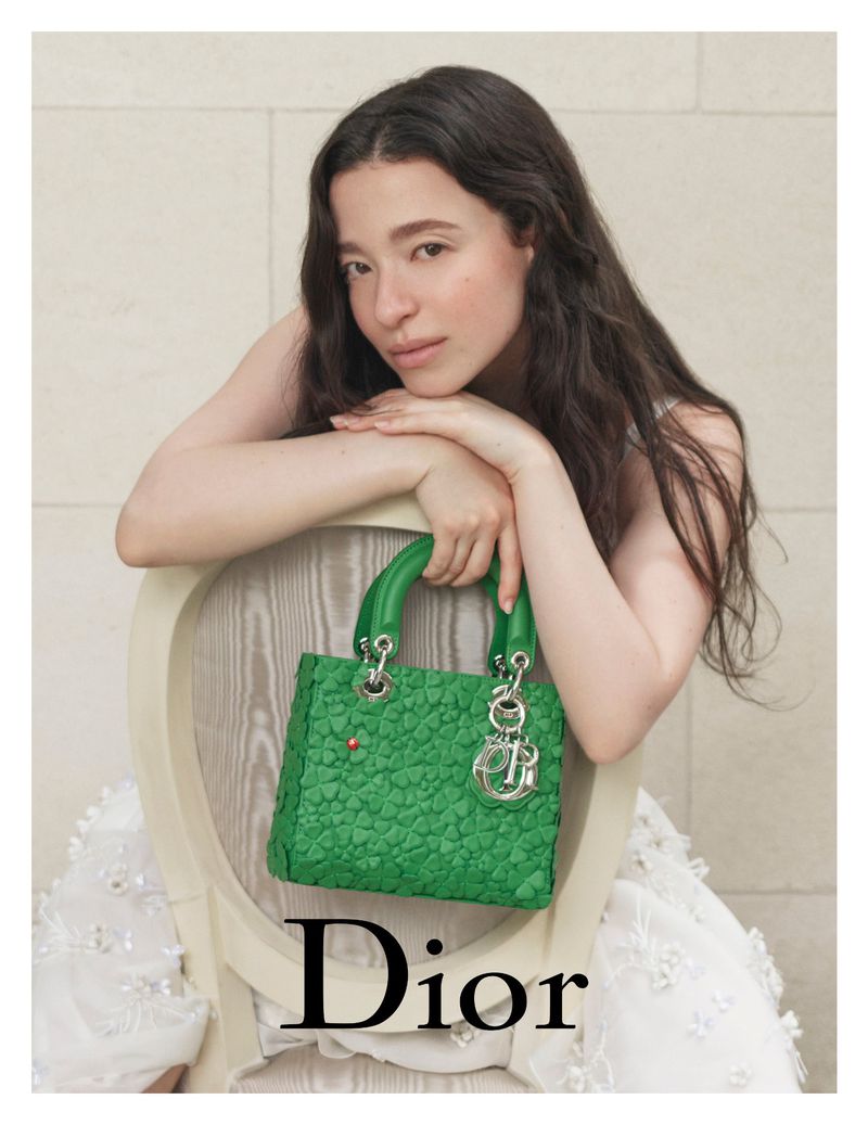 Adult female with long brown hair poses with a green leather Dior handbag with silver hardware against an off-white studio background.
