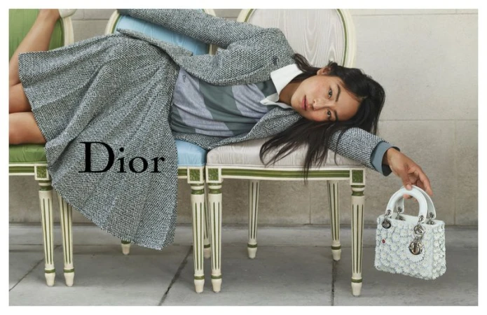 Fashion shot of a woman in a skirt suit reclining on chairs, holding a small decorative purse, with the Dior logo prominently displayed.