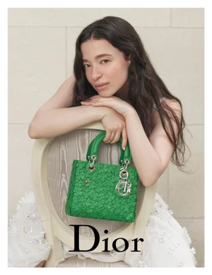 Mikey Madison leaning over an oval-backed chair holding a green textured Lady Dior handbag against a pale stone wall.