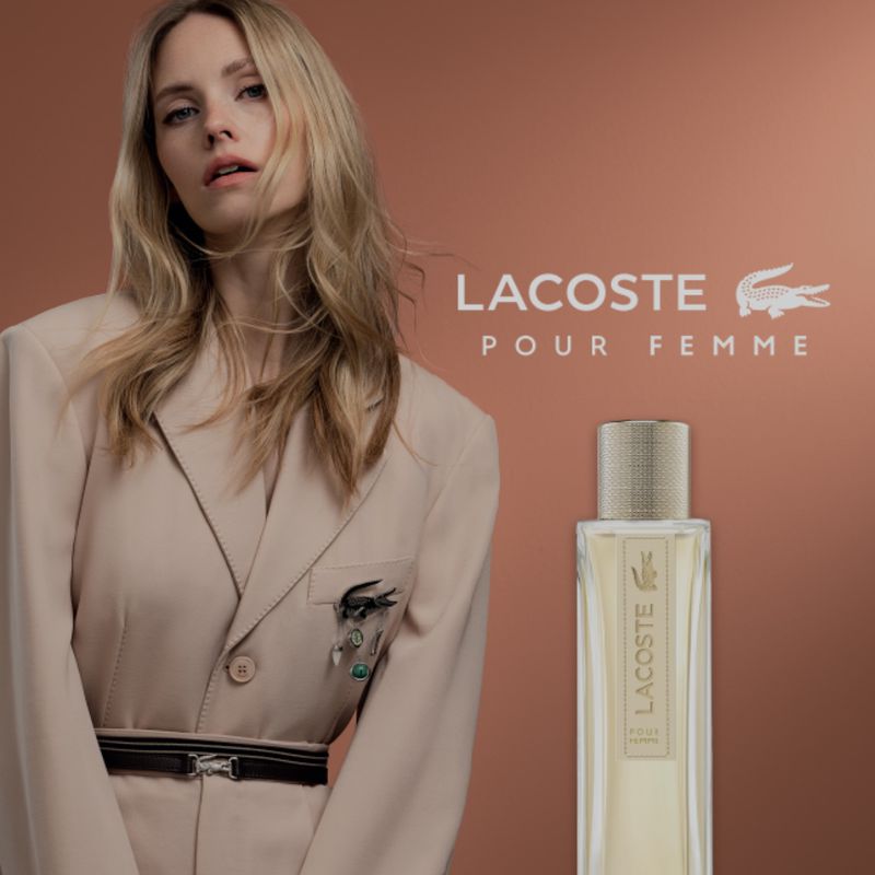 Elegant blonde woman in a beige blazer posing with a bottle of Lacoste Pour Femme perfume against a gradient brown background.