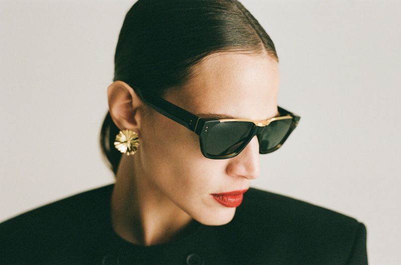 Profile shot of a female fashion model wearing sunglasses, large gold earrings, a dark jacket, and red lipstick, against a light backdrop.