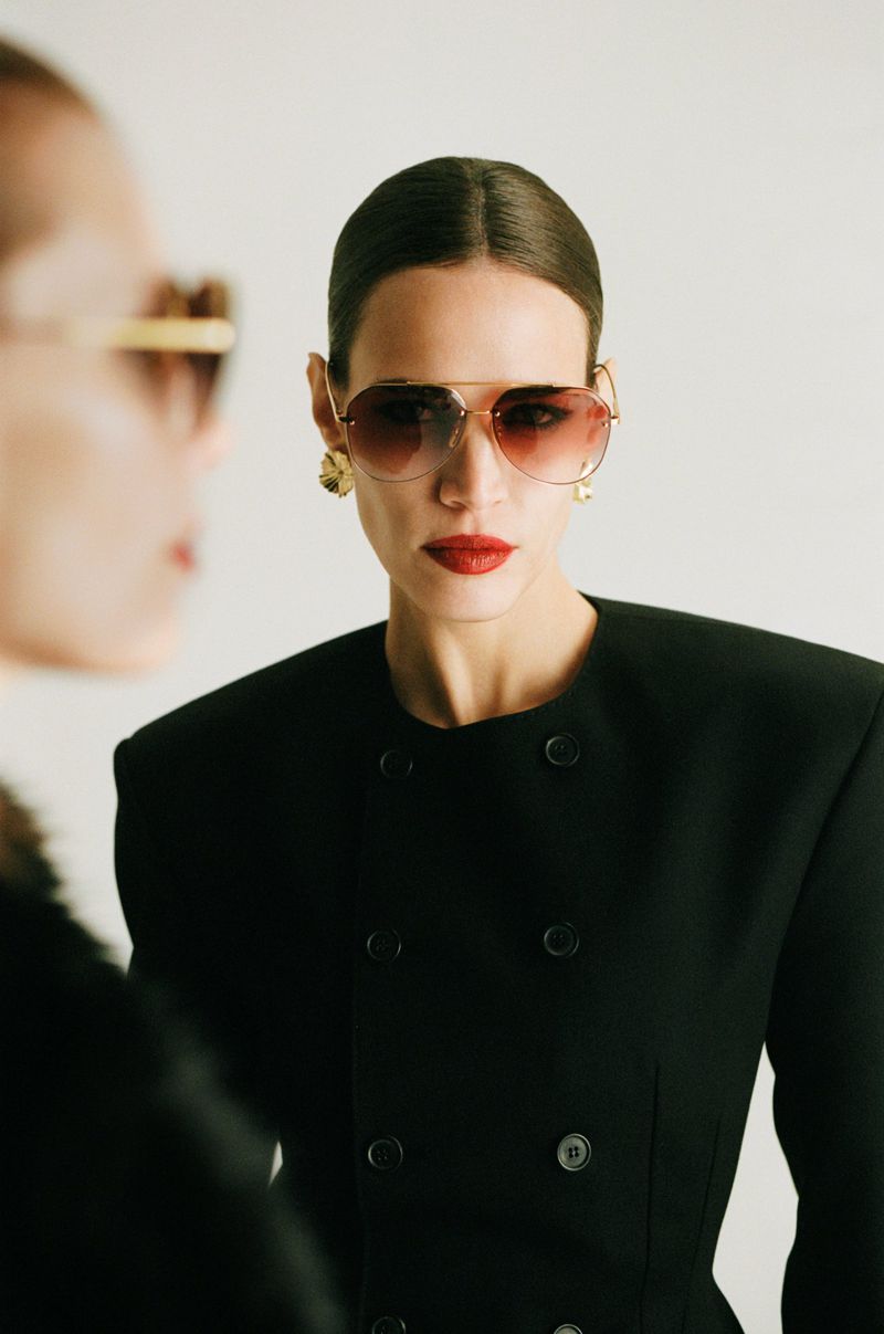 Two adult women pose indoors. One wears sunglasses, a black jacket, and red lipstick. Neutral background, studio lighting, fashion shot.