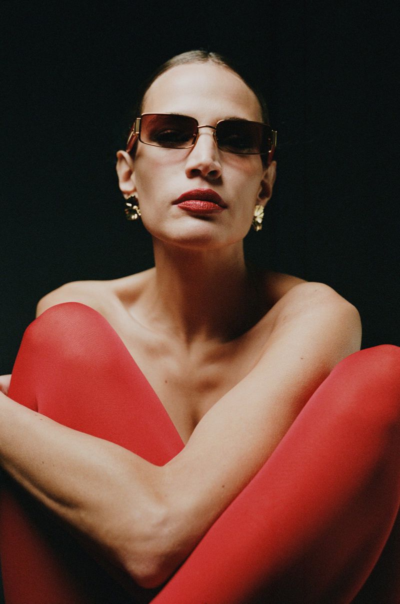 A woman with red lipstick poses in red tights and gold earrings and sunglasses, set against a dark background in a studio shot.