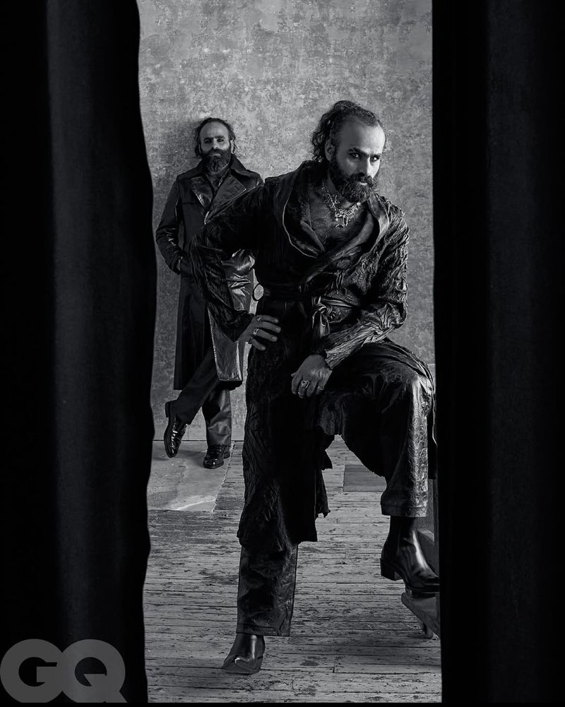 Black-and-white portrait of two bearded adult males in dark clothing against a textured wall, framed by a dark foreground.