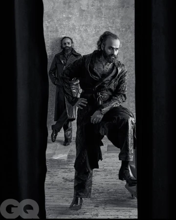 Black-and-white portrait of two bearded adult males in dark clothing against a textured wall, framed by a dark foreground.