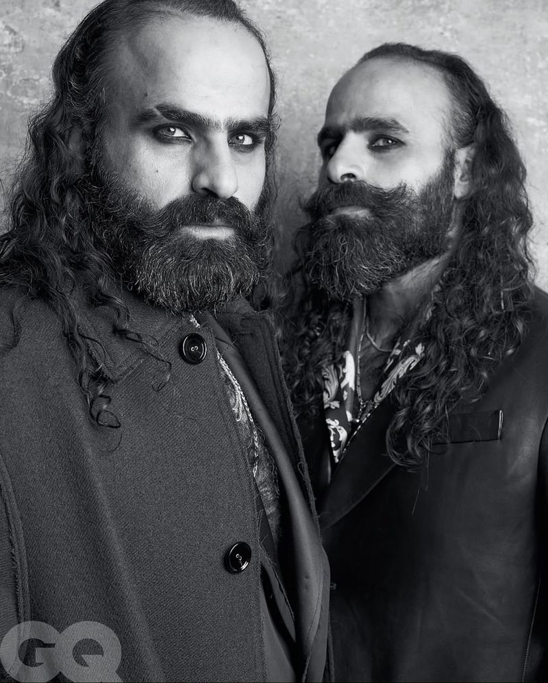 Black and white portrait of two stylish adult males with long hair and beards wearing patterned shirts and dark jackets, close together.