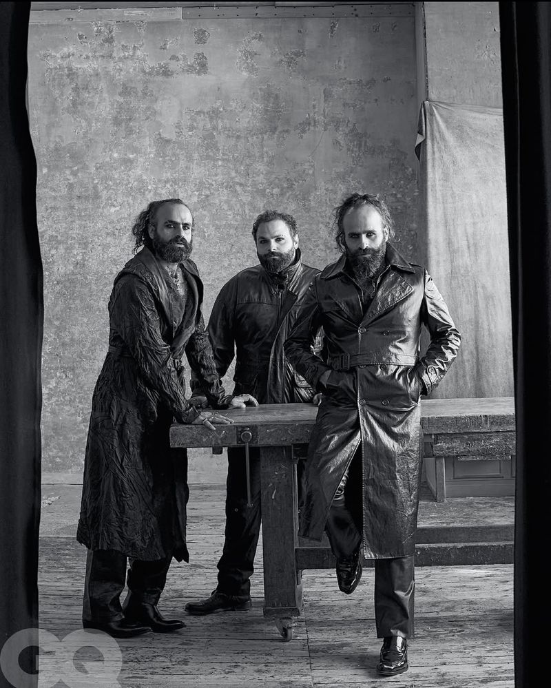 Black and white studio portrait of three adult males with beards wearing dark coats standing behind a workbench with a grunge backdrop.