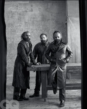 Black and white studio portrait of three adult males with beards wearing dark coats standing behind a workbench with a grunge backdrop.