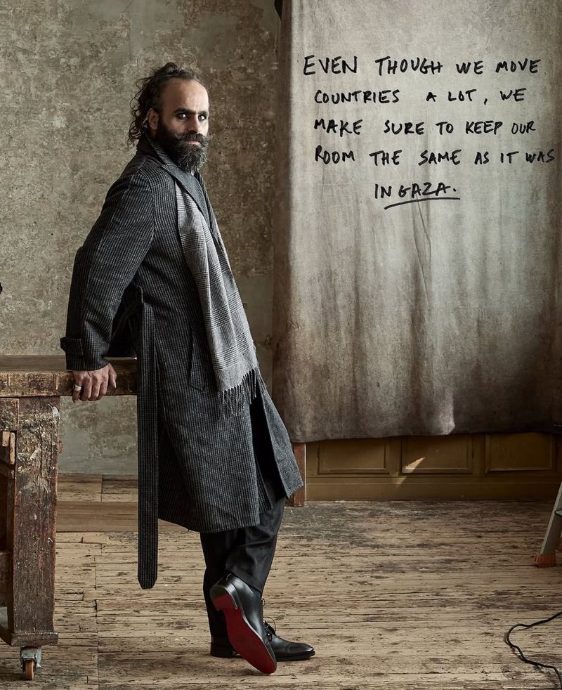 Bearded man in a grey coat poses indoors next to a wooden table. He is standing in front of a canvas with handwritten text on it.