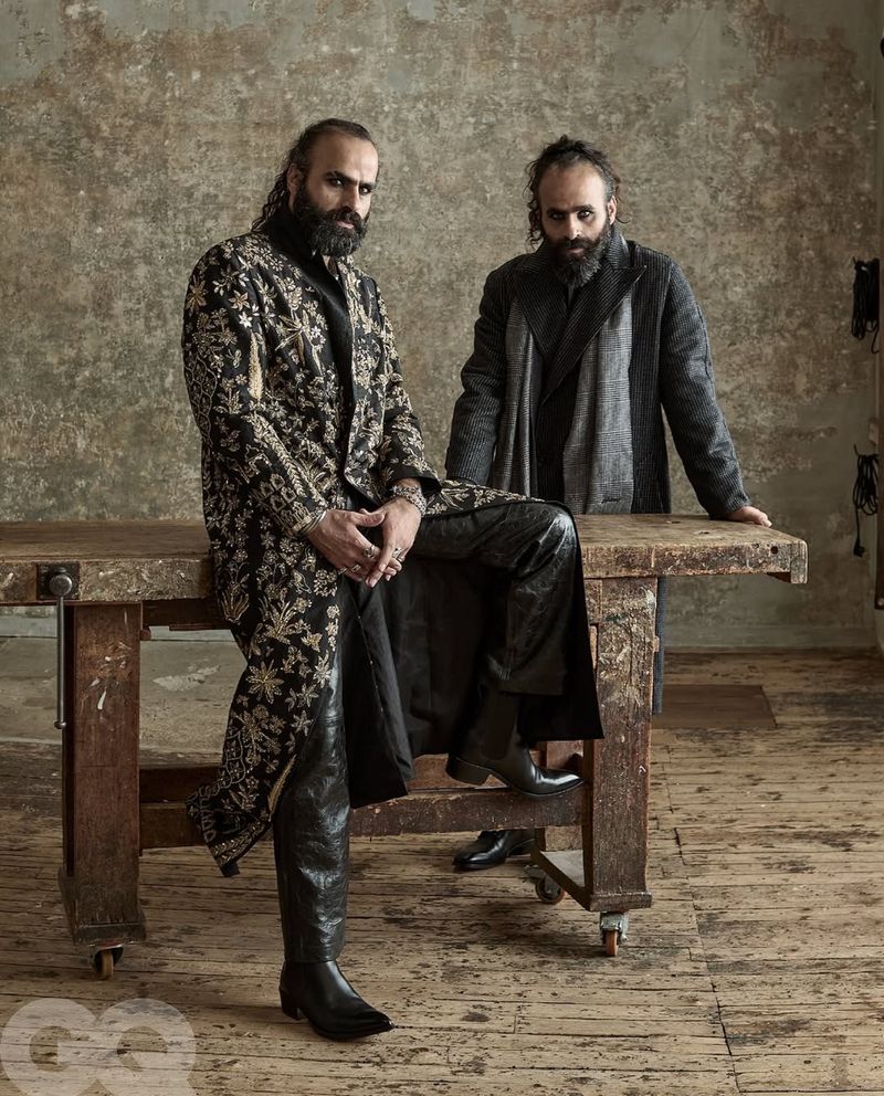 Two bearded men in stylish coats pose indoors against a textured wall, leaning on a wooden table in a studio setting for a fashion editorial.
