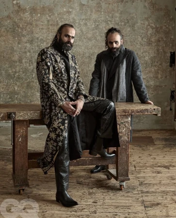 Two bearded men in stylish coats pose indoors against a textured wall, leaning on a wooden table in a studio setting for a fashion editorial.