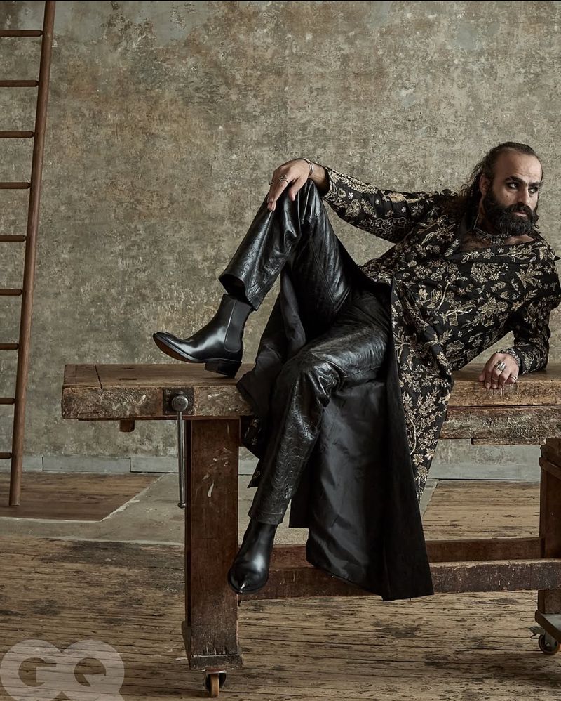 Man with a beard poses in patterned jacket and leather pants, reclining on a wooden table with a ladder and grey textured wall.