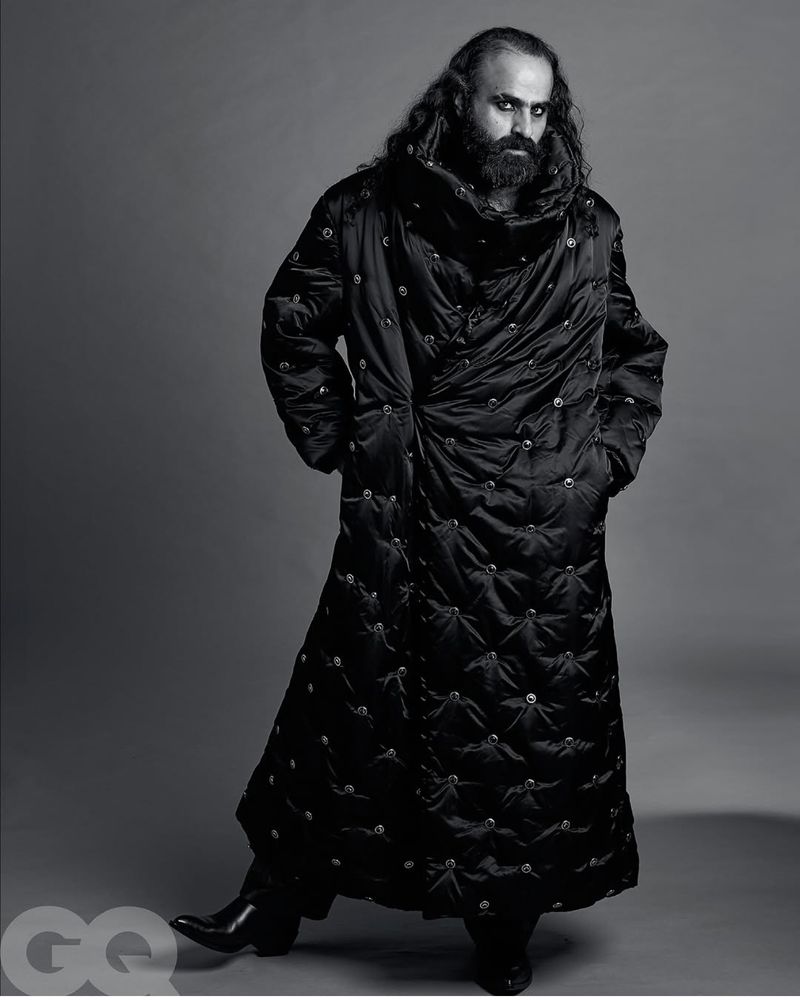 Man in full-length black puffer coat stands in studio with hands in pockets. Fashion editorial photography features dark, moody aesthetic.