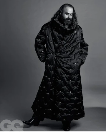 Man in full-length black puffer coat stands in studio with hands in pockets. Fashion editorial photography features dark, moody aesthetic.