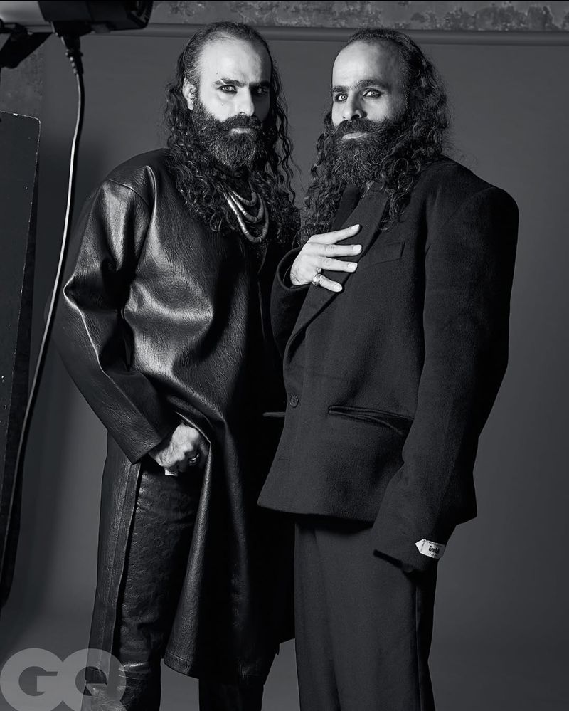 Two men with dark hair and beards stand in a studio, one in leather, one in a suit, in a black and white fashion portrait.