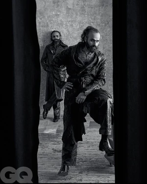 Monochrome portrait of two bearded men posing in textured dark coats against a textured interior wall.