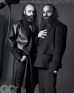 Two men with long dark hair, beards, and heavy eye makeup pose in black tailored garments against a neutral studio backdrop.
