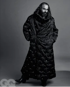 Man with long dark hair and beard wearing a long quilted black coat against a seamless grey backdrop.