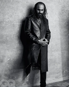 Man with long hair and a beard wearing a long leather coat and pointed boots leaning against a textured wall in black and white.