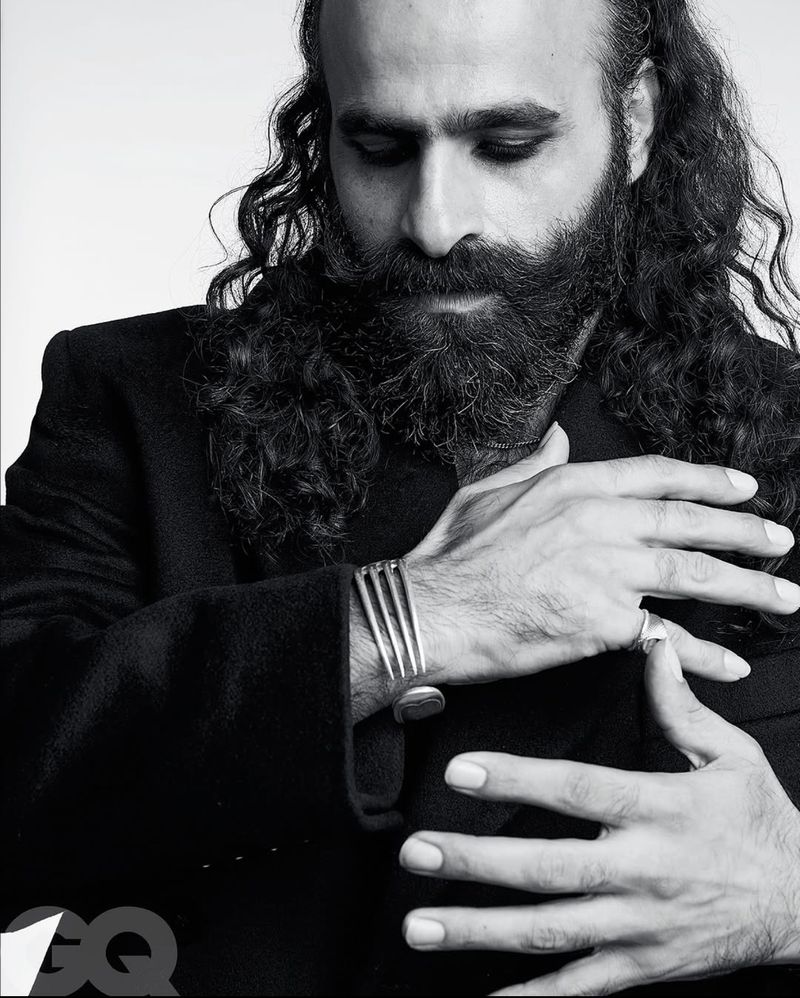 Black and white portrait of a bearded man with long hair wearing a black jacket and a silver cuff bracelet.