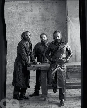 Three bearded men wearing dark long coats and jackets pose around a rustic wooden table in a black-and-white studio setting.