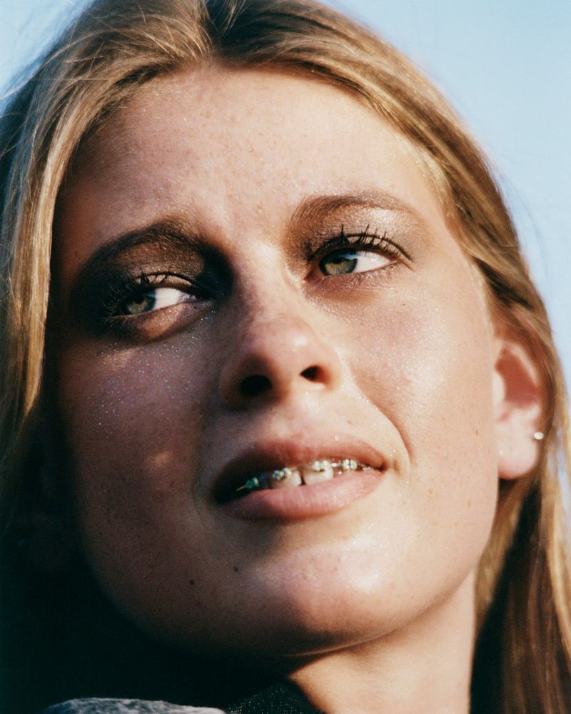 Portrait of a young woman with blond hair and braces smiling slightly, looking upward toward the sky in an outdoor setting.