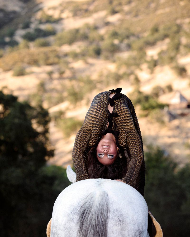 Adult female performs a backbend pose on a gray horse in a sunlit rural setting with tan grassy hills and scattered trees in the background.