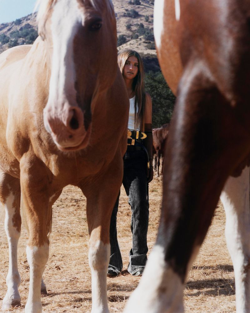 A young woman stands between two tan horses outdoors in a rural setting. The woman wears a white tank and black pants with a wide belt.