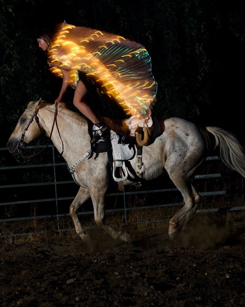 A woman rides a horse at night, wearing a bright, light-up wing-like garment. The horse is mid-gallop in a dark, rural environment.
