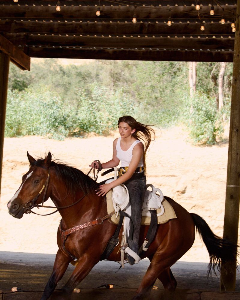A young woman rides a brown horse at an outdoor arena with a wooden structure overhead. The horse and rider are moving to the left.