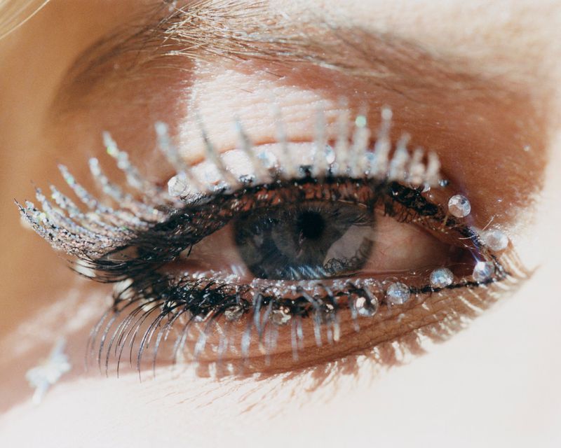 Close-up shot of a woman's eye with dramatic makeup, featuring embellished eyelashes with glitter and rhinestones for a glamorous effect.