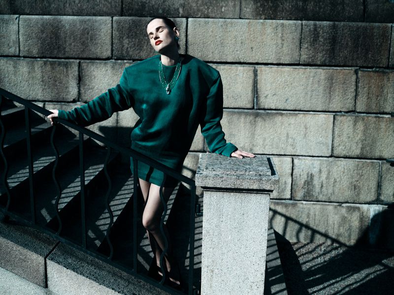 Adult woman poses on stone steps in a short green dress and black shoes, accessorized with a gold necklace and earrings.