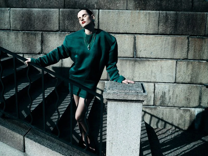 Adult woman poses on stone steps in a short green dress and black shoes, accessorized with a gold necklace and earrings.