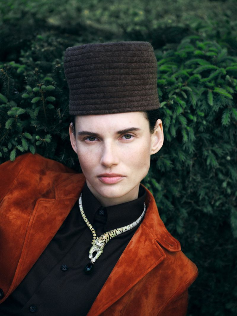 Woman poses in an outdoor portrait with a brown hat, orange jacket, and snake necklace against a green foliage background.
