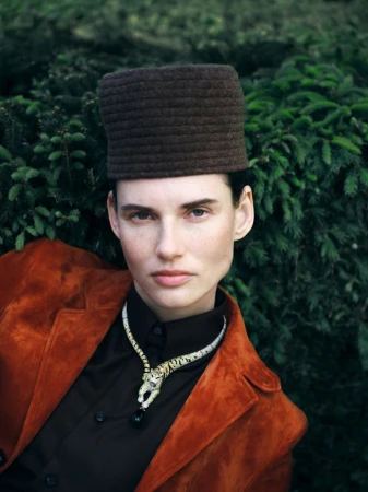 Woman poses in an outdoor portrait with a brown hat, orange jacket, and snake necklace against a green foliage background.